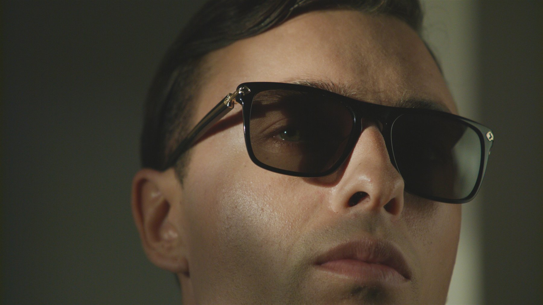 Editorial photograph of a person wearing dark sunglasses in soft shadow.